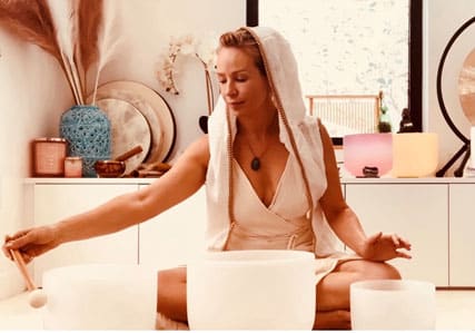 A woman in a light outfit with a hood sits cross-legged indoors, playing crystal singing bowls with a mallet. The background features decorative vases, instruments, and soft natural light.