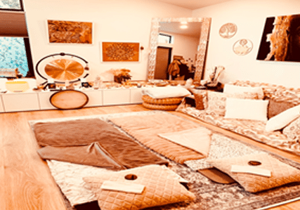 A cozy, bohemian-style room with warm lighting, layered rugs, plush cushions, folded blankets, a large wall mirror, and zen-inspired decor, including a gong and nature-themed artwork on the walls.