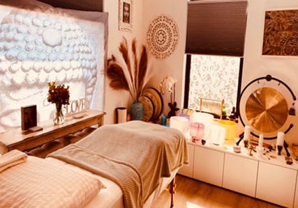 A peaceful massage therapy room with a treatment table, soft blankets, decorative wall art, a Buddha image, dried pampas grass, singing bowls, a gong, and gentle natural light from two windows.