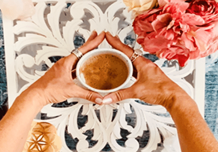 A person with rings on both hands holds a cup of coffee over a white ornate table, with pink flowers and a candle nearby. The scene appears cozy and inviting.