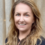 A woman with wavy, light brown hair and blue eyes smiles at the camera. She is wearing a black top and standing in front of a beige curtain background.