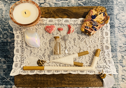 A small altar with a candle, rose quartz crystals, a jar of sand, a shell, a sage bundle with petals, and decorated wands, all arranged on a lace cloth atop a wooden surface.