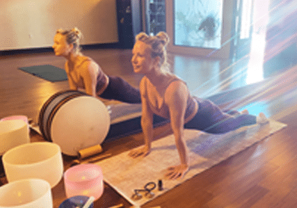 A woman practices yoga indoors on a mat, performing an upward-facing dog pose. She is surrounded by musical bowls and sunlight streams through a nearby window, creating a warm, serene atmosphere.