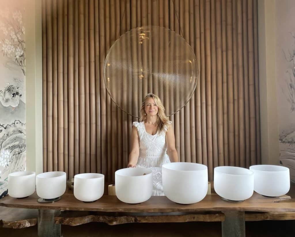 Woman in white dress stands behind a table with seven white crystal singing bowls.