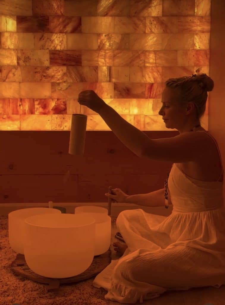 Woman in a white dress plays crystal singing bowls in a warmly lit, amber-colored room.