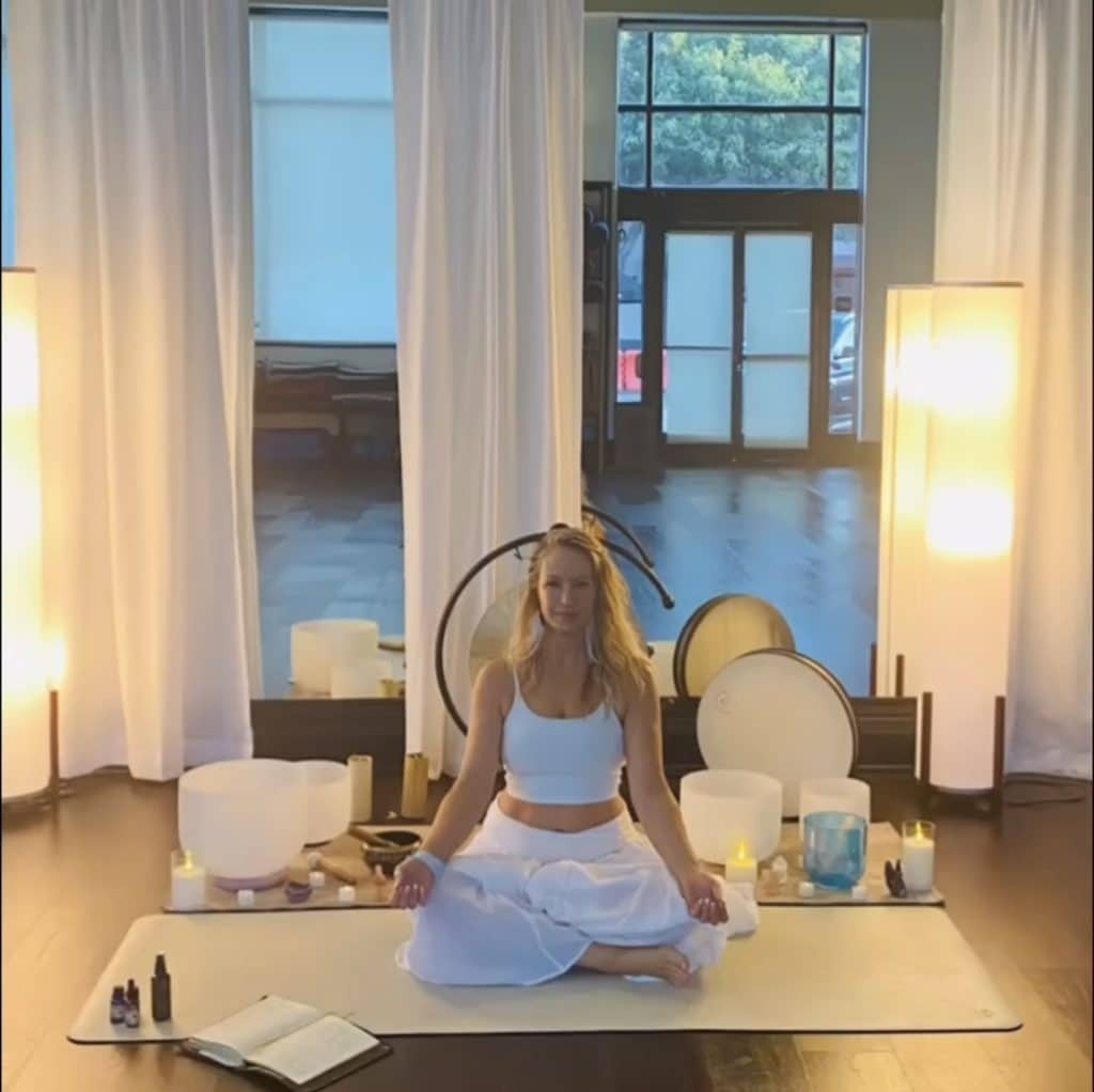 Woman meditating indoors on a mat, surrounded by sound bowls, candles, and an open book.