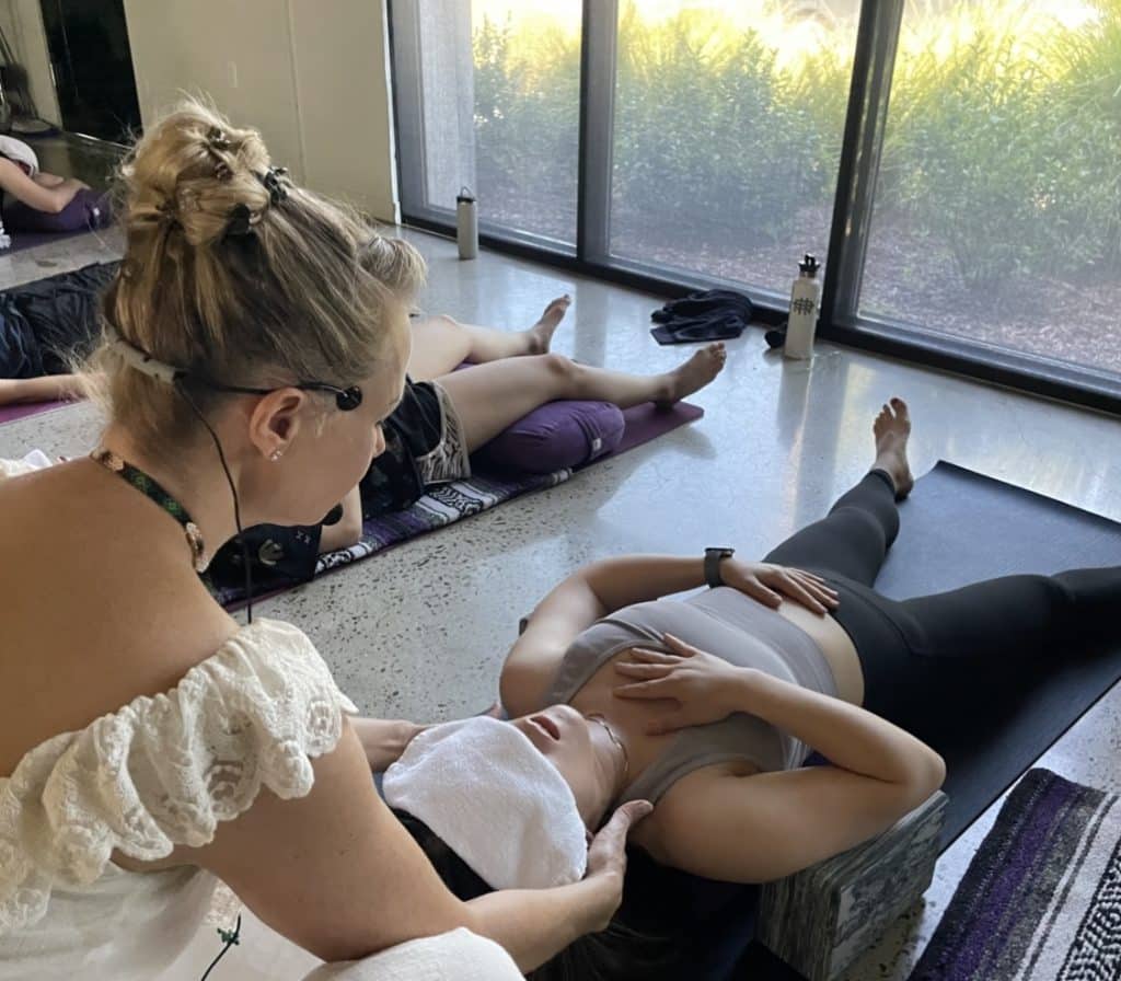 A woman assists another woman lying on a yoga mat with a towel over her eyes.