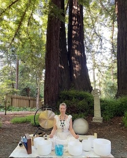 A woman sits among sound bowls outdoors, surrounded by tall redwood trees.